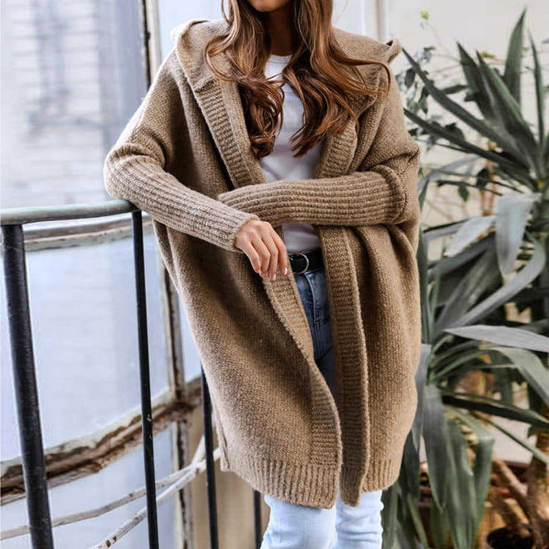 Frau in beigem Strickcardigan, lässig über weißem Shirt und Jeans, steht an einem Fenster. Modetrend, Herbstmode, gemütlicher Look.