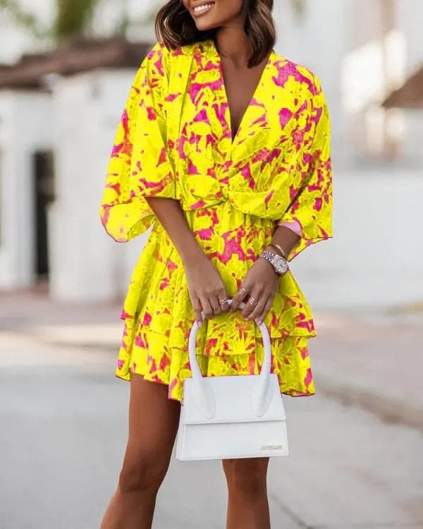 Frau in gelbem, pink gemustertem Kleid mit weißer Handtasche, steht auf Straße. Sommermode, trendiges Outfit, stilvolle Accessoires.