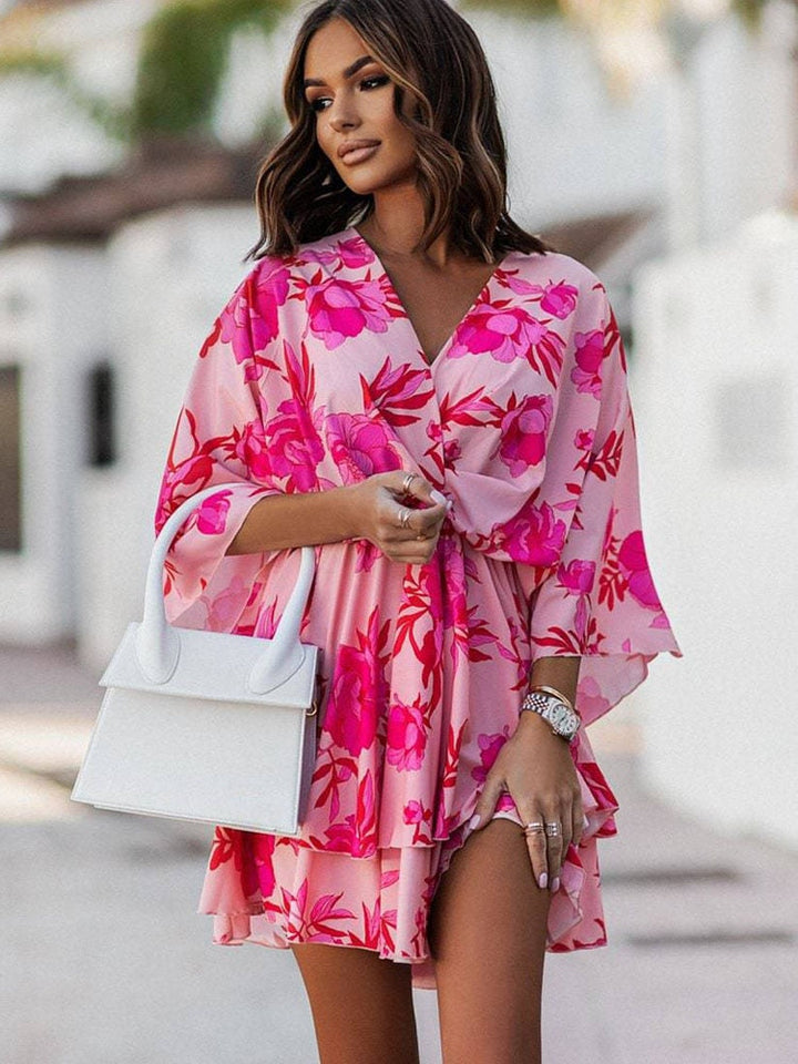 Frau in rosa Blumenkleid mit weißer Handtasche, steht draußen. Sommermode, elegantes Outfit, stilvolle Accessoires, urbane Kulisse.