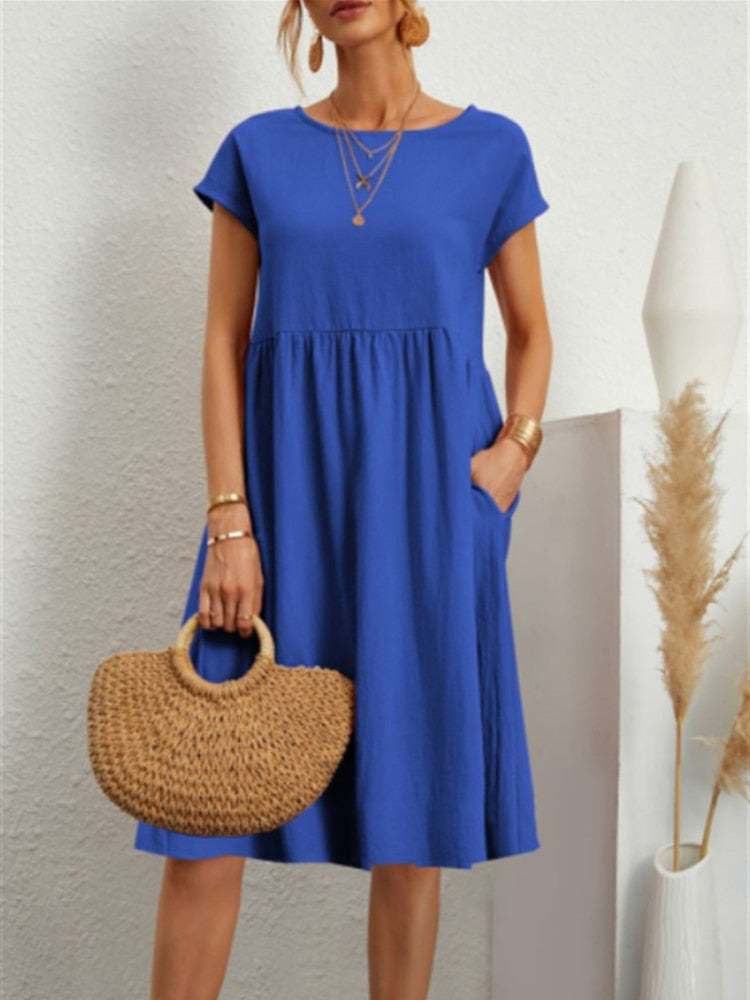 Frau in blauem Sommerkleid mit kurzen Ärmeln, hält eine geflochtene Strohtasche. Minimalistischer Hintergrund mit Pampasgras.