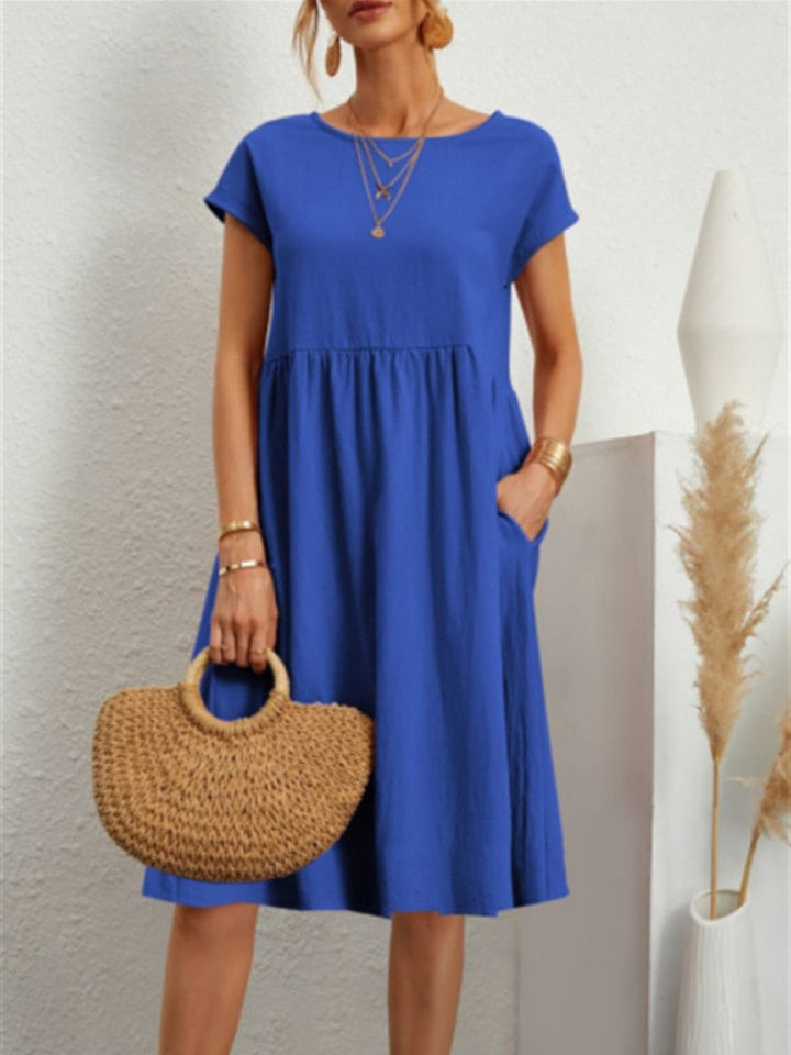 Frau in blauem Sommerkleid mit kurzen Ärmeln, hält eine geflochtene Strohtasche. Minimalistischer Hintergrund mit Pampasgras.