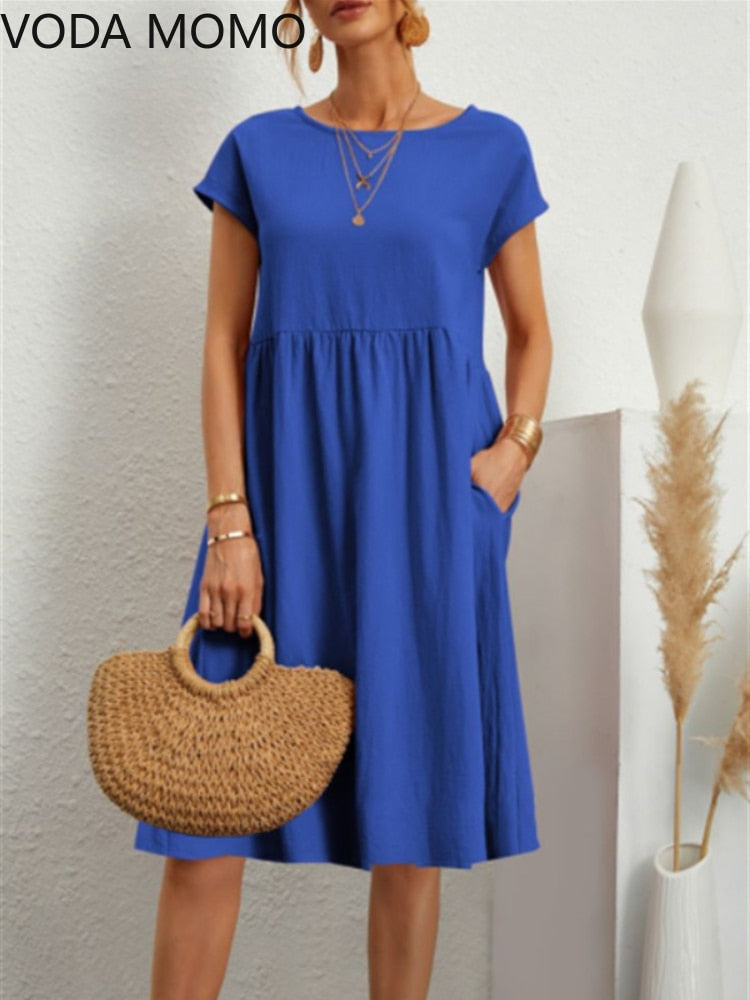 Frau in blauem Sommerkleid mit Strohtasche, steht vor weißer Wand. Mode, Damenbekleidung, Sommeroutfit, Freizeitkleid, Accessoires.