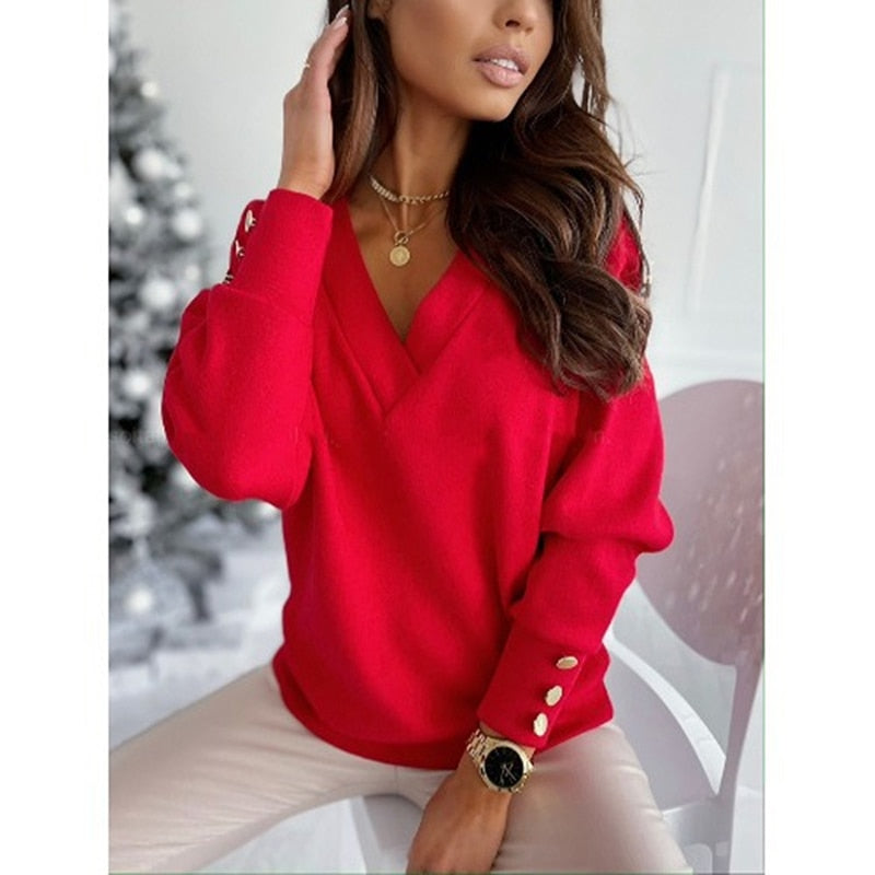 Frau in rotem Pullover mit V-Ausschnitt und goldenen Knöpfen, sitzt auf weißem Stuhl. Weihnachtsbaum im Hintergrund. Elegante Wintermode.