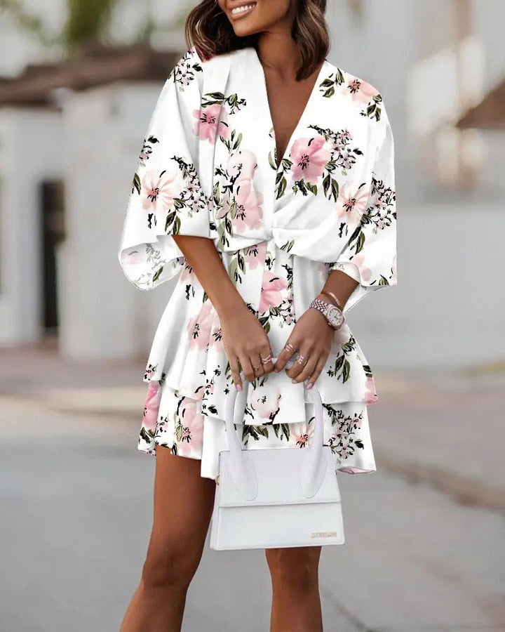 Frau in weißem, geblümtem Sommerkleid mit rosa Blumen, hält weiße Handtasche. Elegante Mode, femininer Stil, trendiges Outfit für Frühling/Sommer.