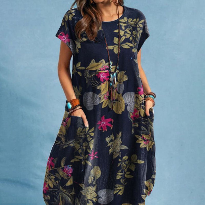 Frau in einem dunkelblauen, geblümten Sommerkleid mit kurzen Ärmeln und Taschen, trägt Armbänder und eine lange Halskette, vor blauem Hintergrund.