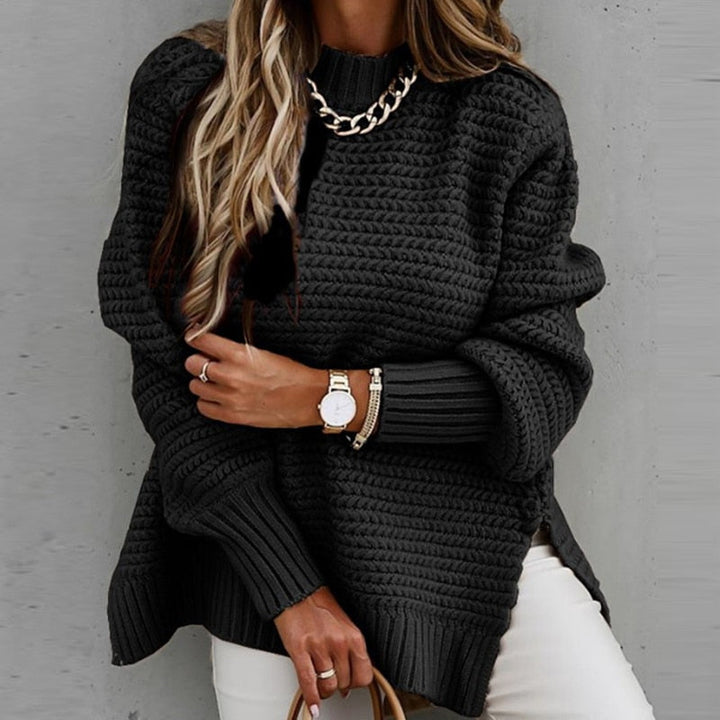 Frau in schwarzem Strickpullover mit Kettendetail, trägt Armbanduhr und Armband, kombiniert mit weißer Hose. Modetrend Herbst/Winter.