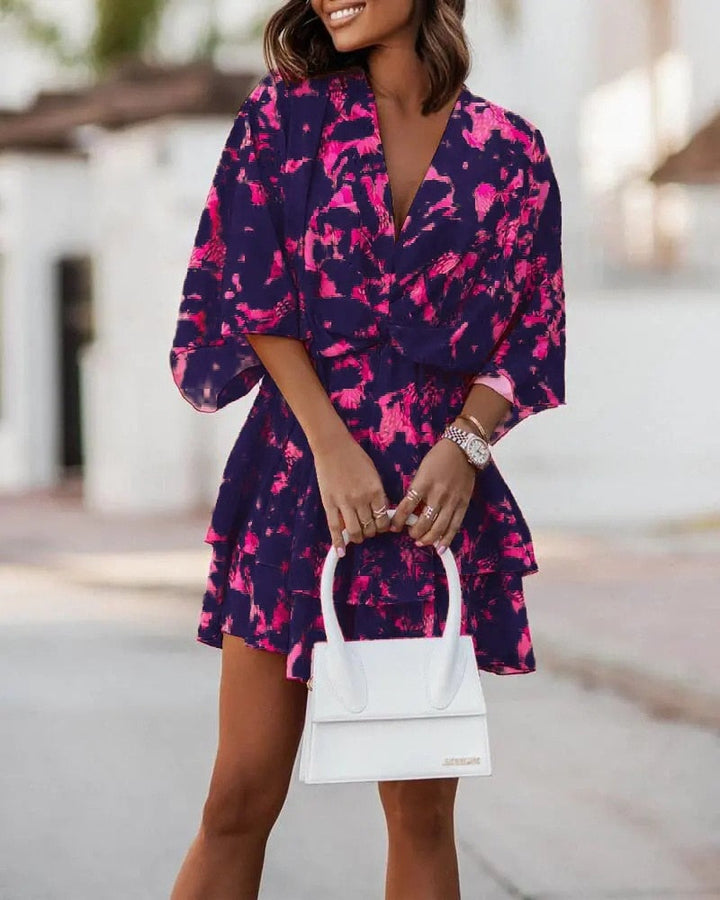 Frau in lila-pinkem Kleid mit abstraktem Muster, hält weiße Handtasche. Sommermode, elegantes Outfit, stilvolle Damenbekleidung.