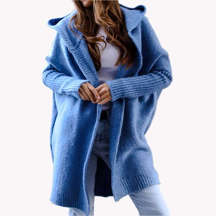 Frau in blauem Strick-Cardigan mit Kapuze, weißem Oberteil und Jeans vor einem weißen Hintergrund. Modischer Herbst-Look, gemütlicher Oversize-Stil.