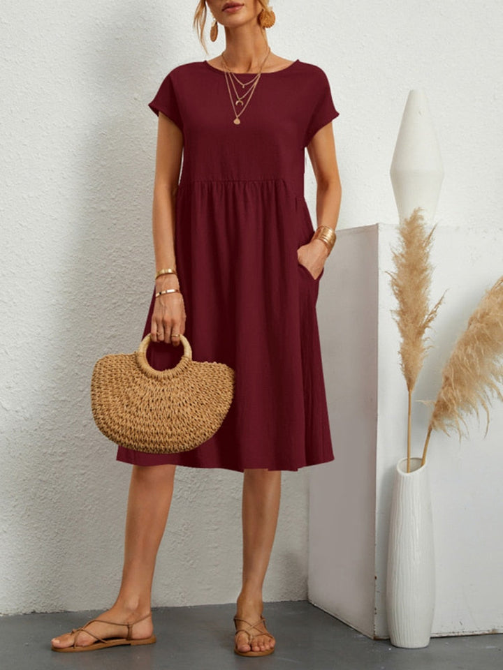 Frau in burgunderrotem Sommerkleid mit Strohtasche, Sandalen und Goldschmuck. Steht neben Vase mit Pampasgras. Elegante, lässige Mode.