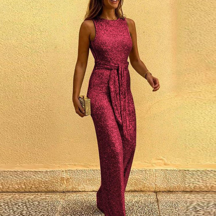 Frau in elegantem, ärmellosem, rotem Paillettenkleid mit Gürtel, hält eine goldene Clutch, steht vor beigem Hintergrund. Abendmode, glamourös.