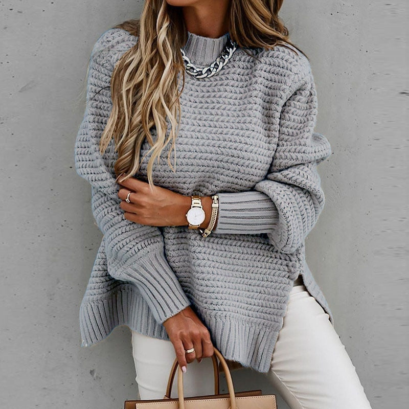 Frau in grauem Strickpullover mit Kettendetail, trägt Armbanduhr und Armbänder, hält braune Handtasche. Modetrend Herbstmode 2023.