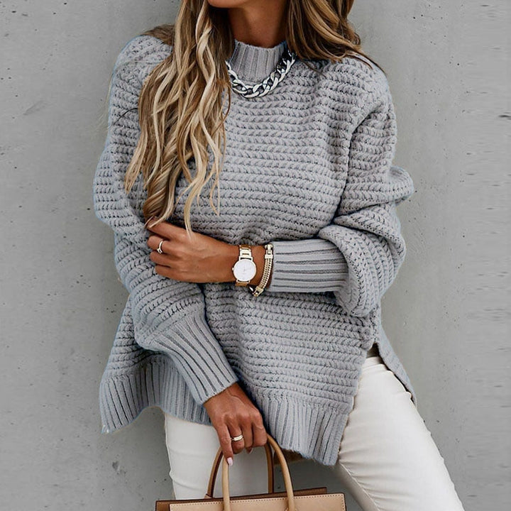 Frau in grauem Strickpullover mit Kettendetail, trägt Armbanduhr und Armbänder, hält braune Handtasche. Modetrend Herbstmode 2023.