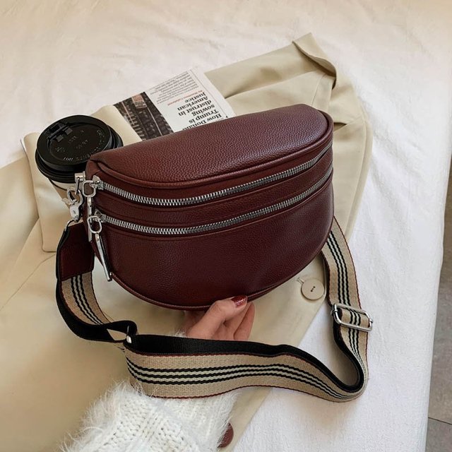 Braune Leder-Bauchtasche mit Doppelreißverschluss und gestreiftem Schultergurt auf beigem Mantel, neben einer Kaffeetasse und Zeitschrift.