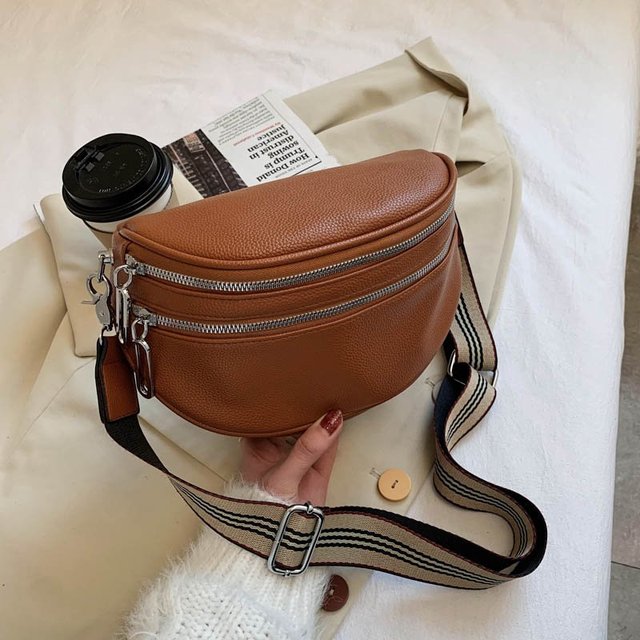 Braune Leder-Gürteltasche mit gestreiftem Schultergurt auf beigem Stoff, neben Kaffeebecher und Buch. Modische Accessoires, stilvoll und praktisch.