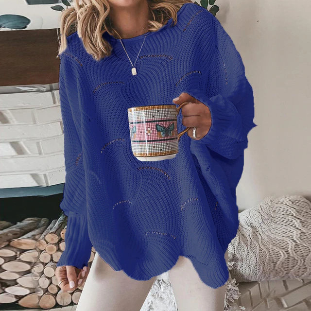Frau in blauem Strickpullover hält Tasse mit Schmetterlingsmotiv, vor Kamin mit Holzstapel. Gemütliche Wintermode, Wohnzimmerdekor.