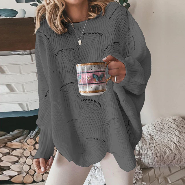 Frau in grauem Strickpullover hält Tasse mit Schmetterlingsmotiv vor Kamin. Gemütliche Wintermode, Strickmode, Kaffeepause.