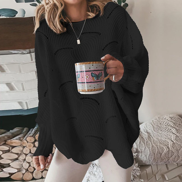 Frau in schwarzem Strickpullover hält Tasse mit Schmetterlingsmotiv, steht vor Kamin. Gemütliche Herbstmode, lässiger Stil, Innenraum.