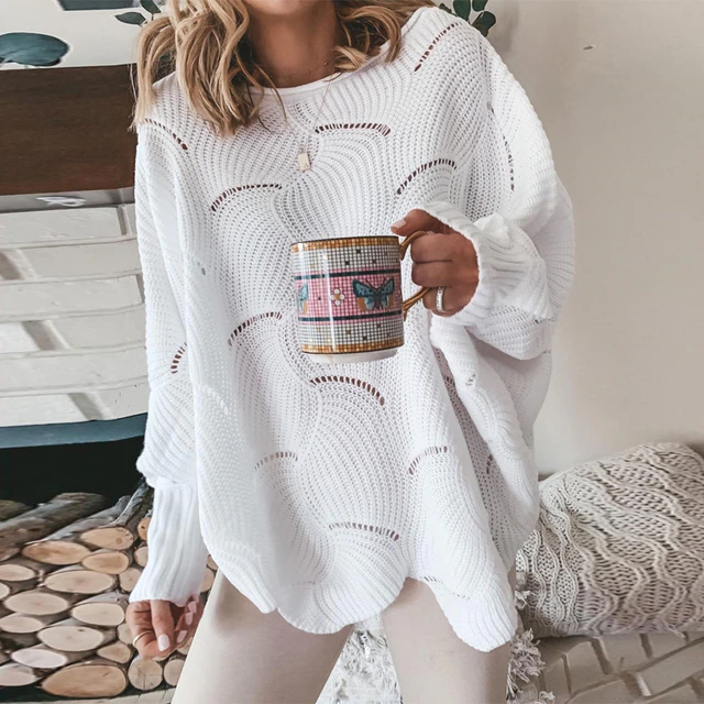 Frau in weißem Strickpullover hält Tasse mit Schmetterlingsmotiv. Gemütliches Wohnzimmer, Kamin im Hintergrund. Herbstmode, Hygge-Stil.