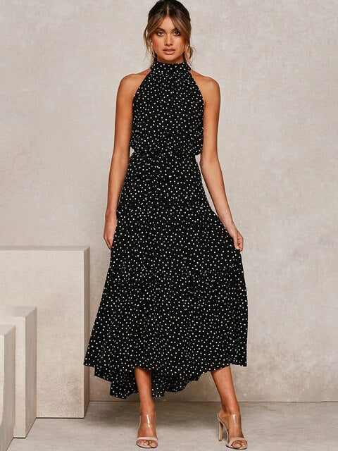 Frau in schwarzem, ärmellosem Kleid mit weißen Punkten, stehend vor neutralem Hintergrund. Elegantes, modisches Sommeroutfit, ideal für Abendveranstaltungen.