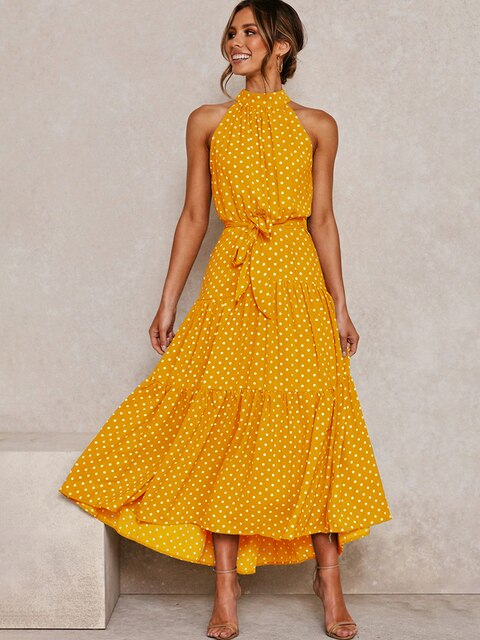Frau in gelbem, gepunktetem Sommerkleid mit Halterneck und Schleife, lächelnd, stehend vor neutralem Hintergrund. Elegantes, modisches Damenoutfit.
