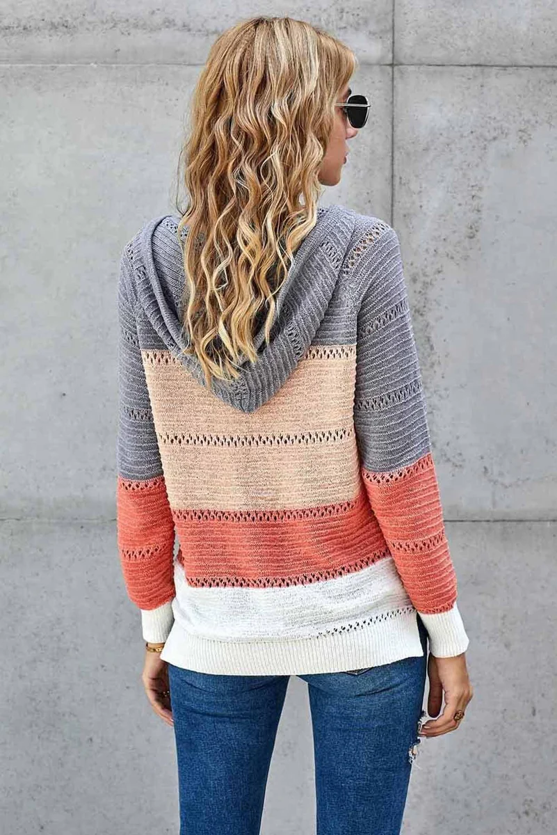 Frau mit lockigem Haar trägt gestreiften Strickpullover in Grau, Beige und Koralle, kombiniert mit blauer Jeans. Modetrend Herbst 2023.