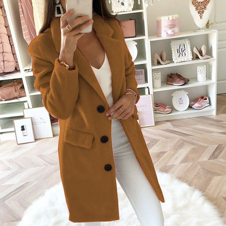 Frau im modischen, senffarbenen Mantel, weißem Top und Hose, macht Selfie in stilvollem Ankleidezimmer mit Regalen und Dekorationen.