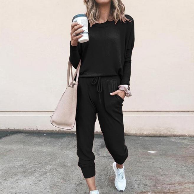 Frau in schwarzem Freizeitanzug mit Kaffeebecher und beiger Handtasche, lässiger Streetstyle, modische Sneakers, urbaner Hintergrund.