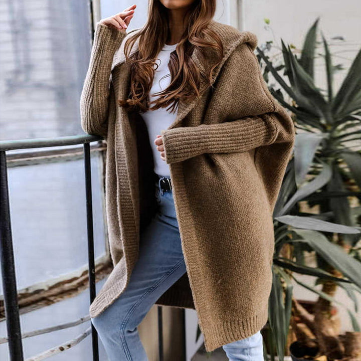 Frau in braunem Strickmantel und blauer Jeans steht auf Balkon, moderne Herbstmode, gemütlicher Oversize-Look, urbaner Hintergrund, Pflanzen.