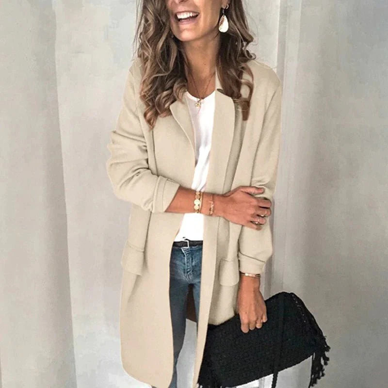 Frau in beigem Mantel, weißem Oberteil und blauer Jeans, hält schwarze Tasche. Modischer Herbst-Look, lässig-elegant, ideal für kühle Tage.