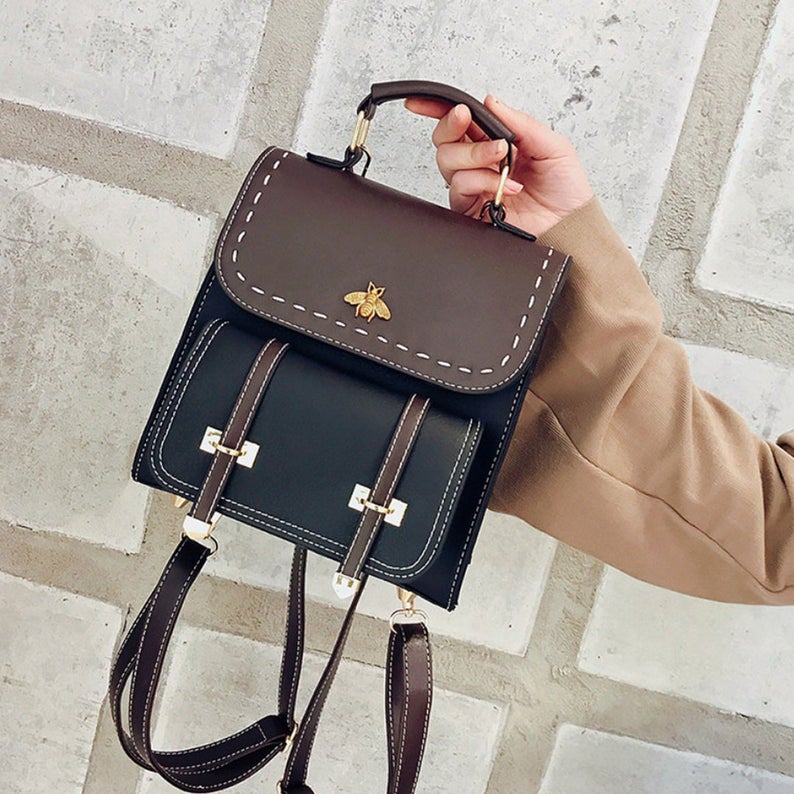 Braune und schwarze Ledertasche mit Bienenmotiv, handgehalten vor grauer Wand. Modische Damenhandtasche, elegantes Design, trendiges Accessoire.