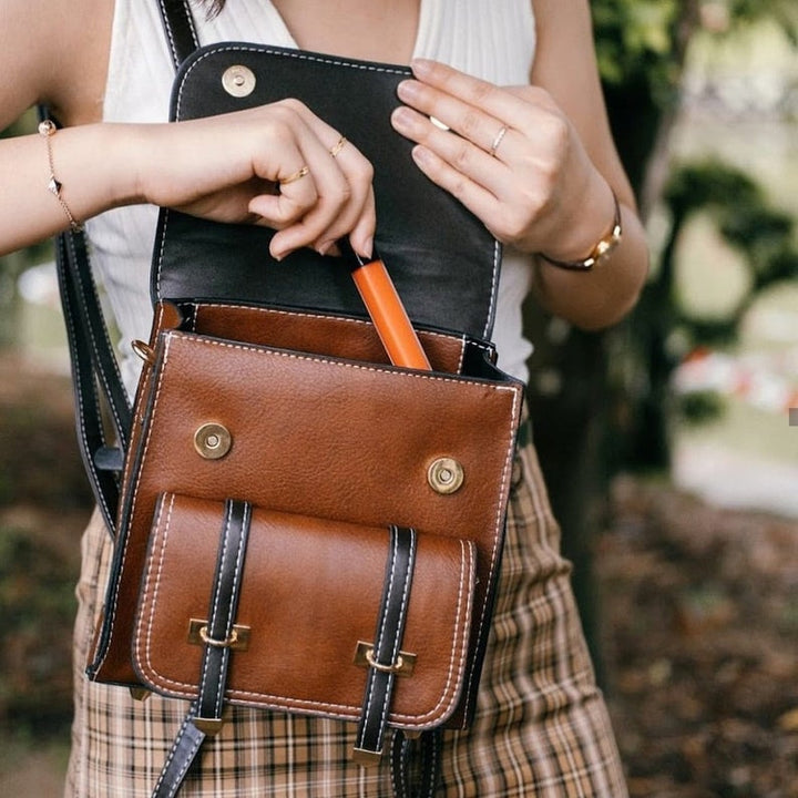 Frau öffnet braune Ledertasche, entnimmt orangefarbenen Gegenstand. Modische Handtasche, Accessoire, Outdoor, stilvoll, Freizeitkleidung.