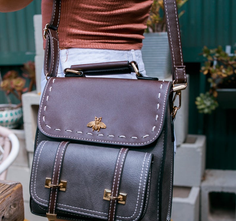 Braune Ledertasche mit Bienenemblem, verstellbarem Schulterriemen und goldenen Schnallen, getragen von Person. Trendiges Accessoire für Damen.