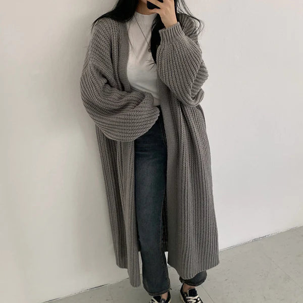 Frau in grauem Strick-Cardigan, weißem T-Shirt und Jeans posiert vor weißer Wand. Modetrend, lässiger Look, Herbstmode, Oversize-Cardigan.