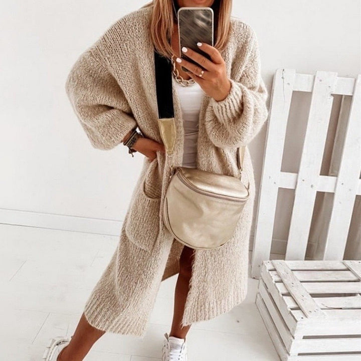 Frau in beigem Strick-Cardigan, weißem Top und Sneakers, mit beiger Umhängetasche, macht Selfie vor weißem Hintergrund. Modetrend Herbst 2023.