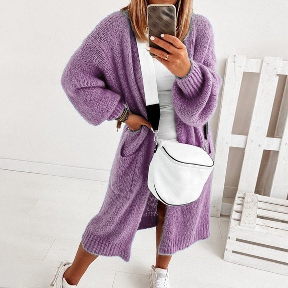 Frau in lila Strickcardigan, weißem Top und Sneakers, hält Smartphone, trägt weiße Umhängetasche. Moderne Herbstmode, lässiger Stil.
