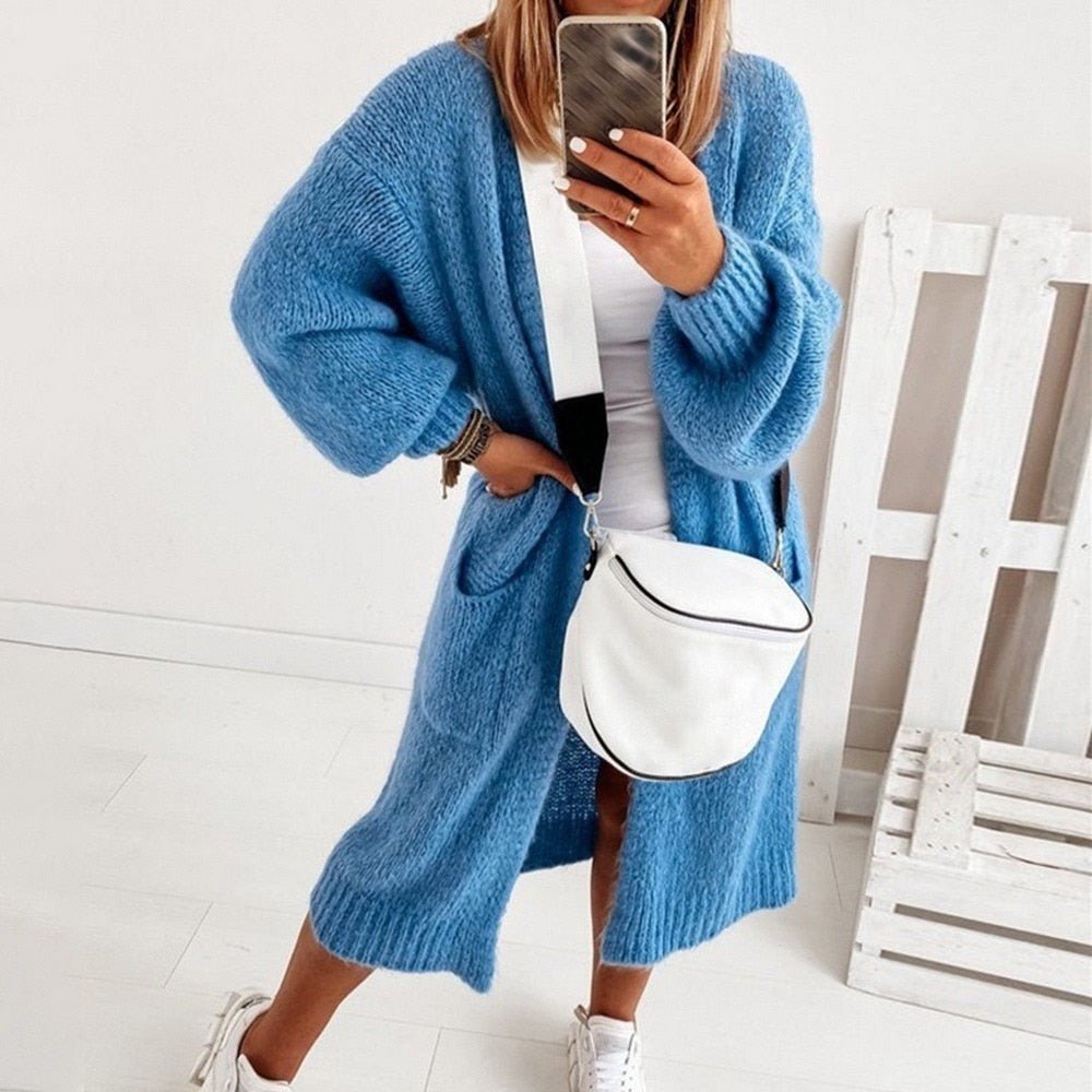 Frau in blauem Strick-Cardigan, weißem Top und Sneakern, hält Smartphone, trägt weiße Umhängetasche. Modetrend, lässiger Herbstlook, Streetstyle.