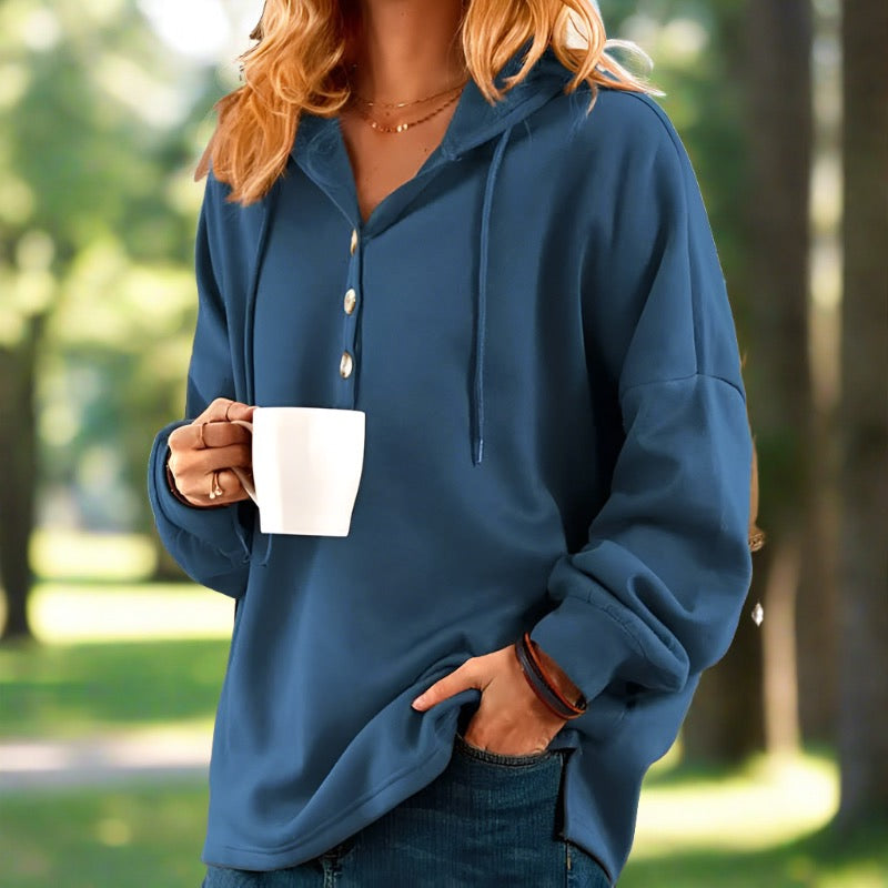Frau in blauem Hoodie hält weiße Tasse, steht im Park. Freizeitmode, Herbst, Kaffeepause, entspannte Atmosphäre, Naturhintergrund.