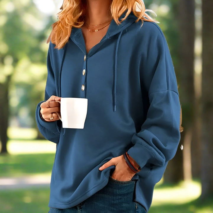 Frau in blauem Hoodie hält weiße Tasse, steht im Park. Freizeitmode, Herbst, Kaffeepause, entspannte Atmosphäre, Naturhintergrund.