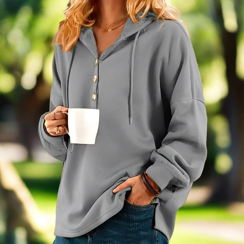 Frau in grauem Hoodie mit Kapuze hält weiße Tasse, lässige Freizeitmode, draußen im Park, entspannte Atmosphäre, Herbstmode.