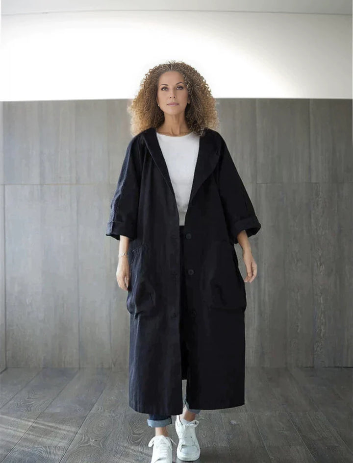 Frau in langem, schwarzem Mantel und weißen Sneakers vor grauer Wand. Modetrend, lässiger Stil, Herbstmode, Damenbekleidung.