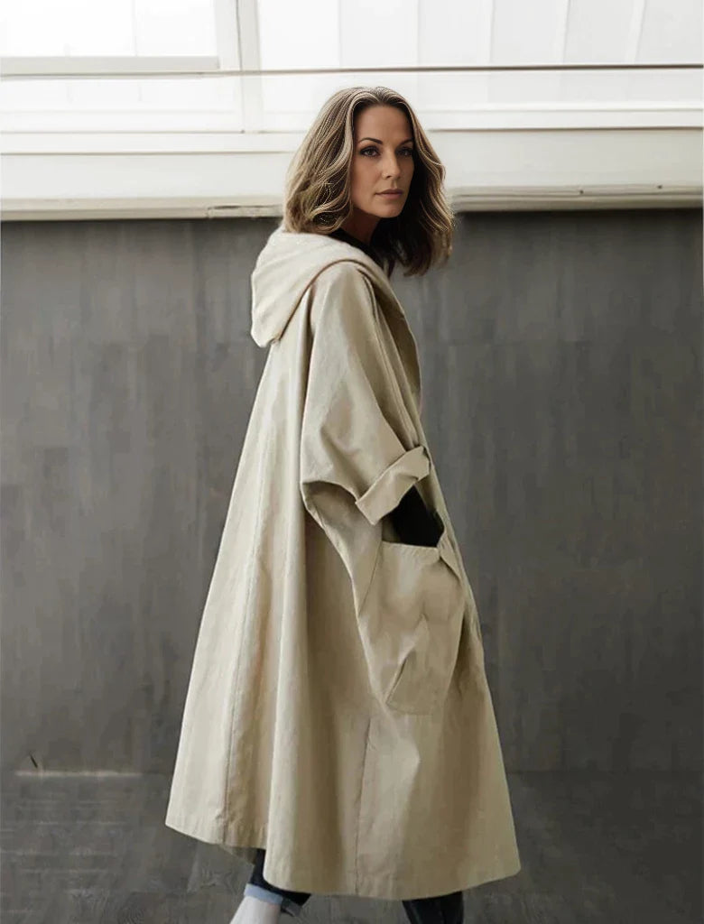 Frau in beigem Oversize-Mantel mit Kapuze, steht vor grauer Wand. Modetrend, Herbstmode, lässiger Stil, Damenbekleidung, urbaner Look.