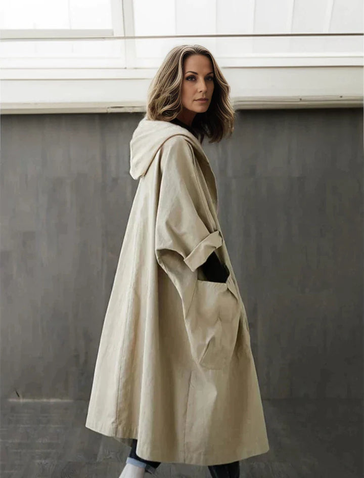 Frau in beigem Oversize-Mantel mit Kapuze, steht vor grauer Wand. Modetrend, Herbstmode, lässiger Stil, Damenbekleidung, urbaner Look.
