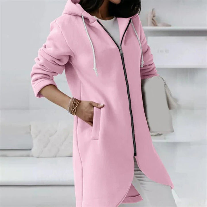 Frau in rosa Kapuzenjacke mit Reißverschluss und Taschen, lässiger Stil, modische Damenbekleidung, gemütliches Wohnzimmer im Hintergrund.