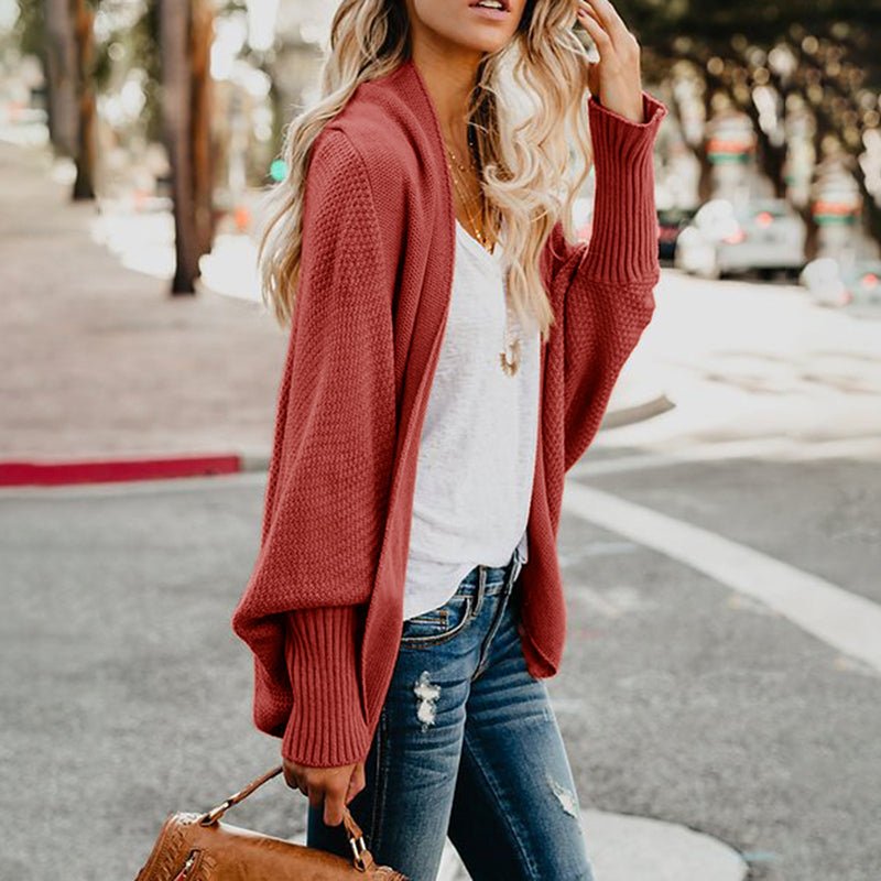 Blonde Frau in rotem Strickcardigan und weißem T-Shirt, trägt Jeans und braune Handtasche, steht auf städtischer Straße. Modetrend Herbst 2023.