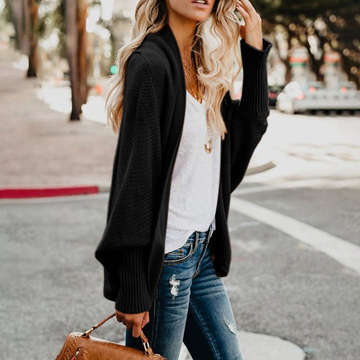 Frau in schwarzer Strickjacke und blauer Jeans, hält braune Handtasche, steht auf Gehweg. Modetrend, Herbstmode, lässiger Stil.